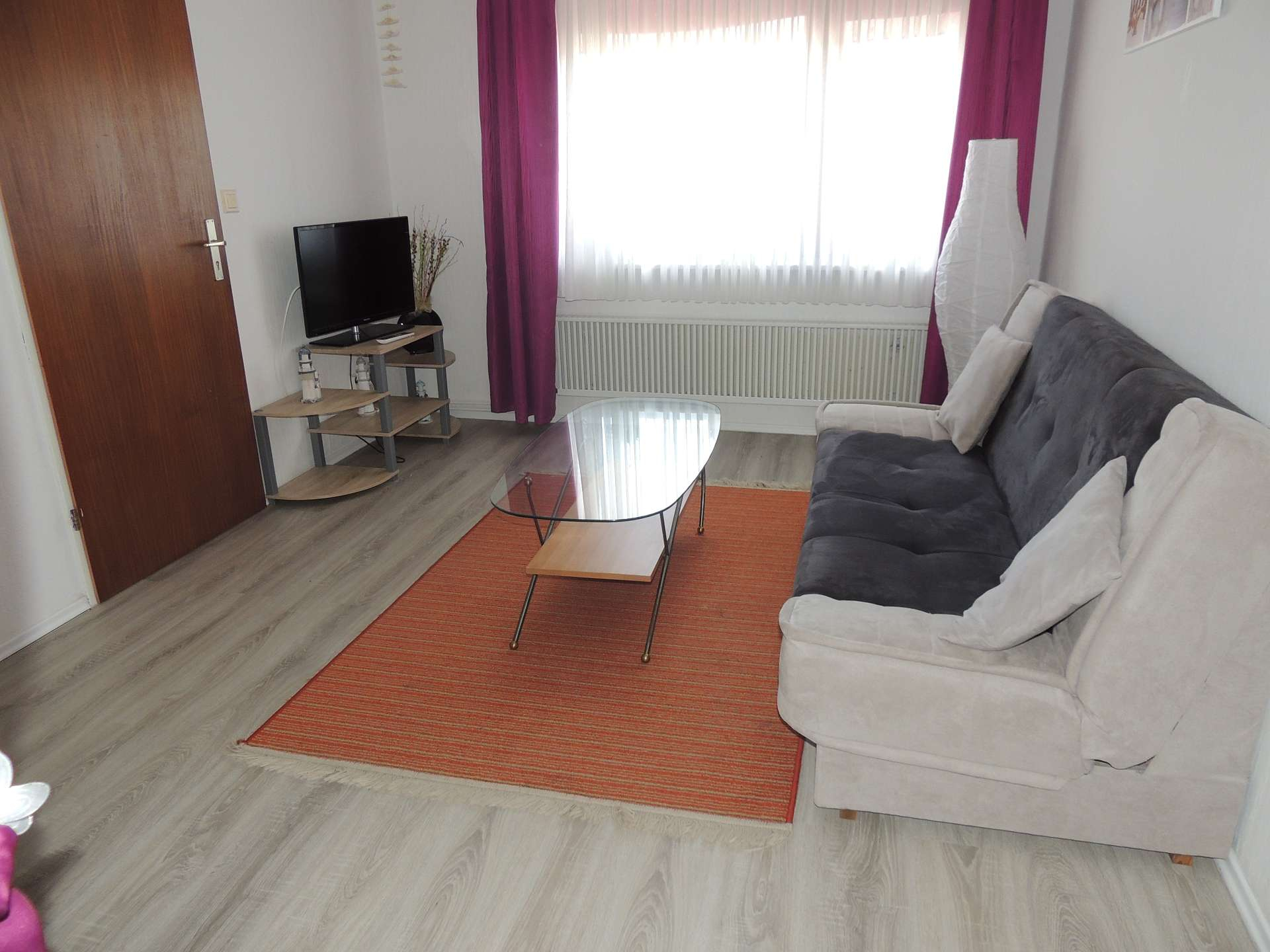 Wohnzimmer mit beige-grauem Sofa, gläsernem Couchtisch auf orangenen Teppich und Fernseher